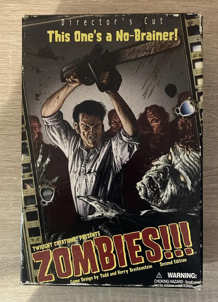 A worn box cover for the "Director's Cut" second edition of the board game "ZOMBIES!!!" featuring an illustration of a man wielding a chainsaw above his head, surrounded by zombies, with the tagline "This One's a No-Brainer!"