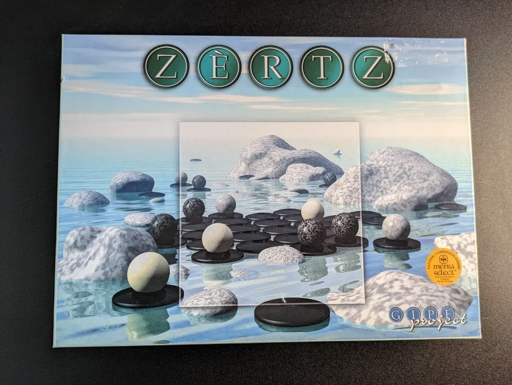 The image shows a "ZERTZ" board game box with a serene sea landscape in the background, featuring rocks in water and black and white gaming pieces on the front. There's a Mensa Select seal on the bottom right and the GIPF project logo at the bottom center.