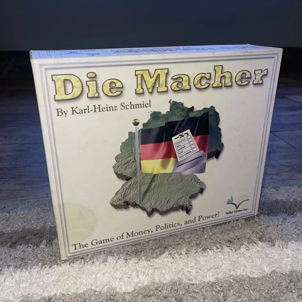 A board game called "Die Macher" by Karl-Heinz Schmiel, featuring a graphic of Germany with a flag and a calculator, titled "The Game of Money, Politics, and Power", produced by Valley Games Inc., sitting on a carpeted floor.