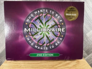 A "Who Wants to Be a Millionaire" 2nd edition board game box on a wooden surface, with a purple background and decorative elements, and a gold sticker reading "UK's Number 1 Board Game 1999, 2000, 2001".