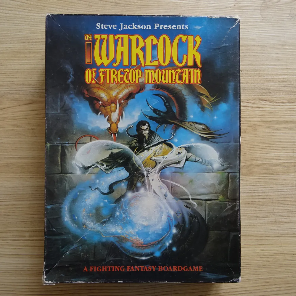 A worn-out board game box with the title "Steve Jackson Presents The Warlock of Firetop Mountain" featuring fantasy artwork including a warlock wielding magic, serpentine creatures, and mystical elements, laid on a wooden surface.