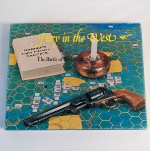 A board game titled "Fury in the West" depicting the Battle of Shiloh, April 6 and 7, 1862, with a book on infantry tactics, an antique revolver, and a brass candle holder on top.