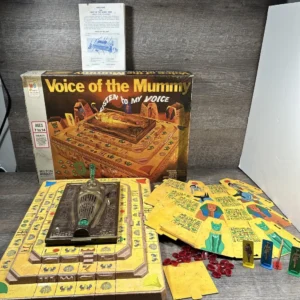 Board game "Voice of the Mummy" by Milton Bradley on a table with its box, instructions, game board, cards, plastic figures, and red tokens displayed.