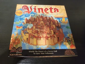 A board game titled "Vineta: Atlantis of the North" is displayed on a dark surface. The cover art features a colorful illustration of a mythical city with buildings in shades of red and yellow, a blue ocean with a sailing ship, and Norse warriors in the foreground. The game's tagline "Wield The Power of a Norse God To Save Your Followers" and the logos for "Immortal Eyes Games" and "Play Forever" are also visible.