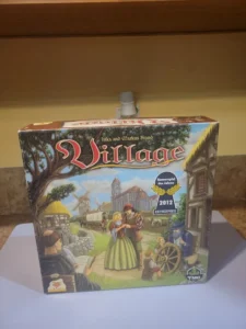 A board game box titled "Village" by Inka and Markus Brand, displayed on a shelf. The box features vivid artwork of medieval village life with characters engaged in various activities and indicates a "2012 Kennerspiel des Jahres Kritikerpreis" award.