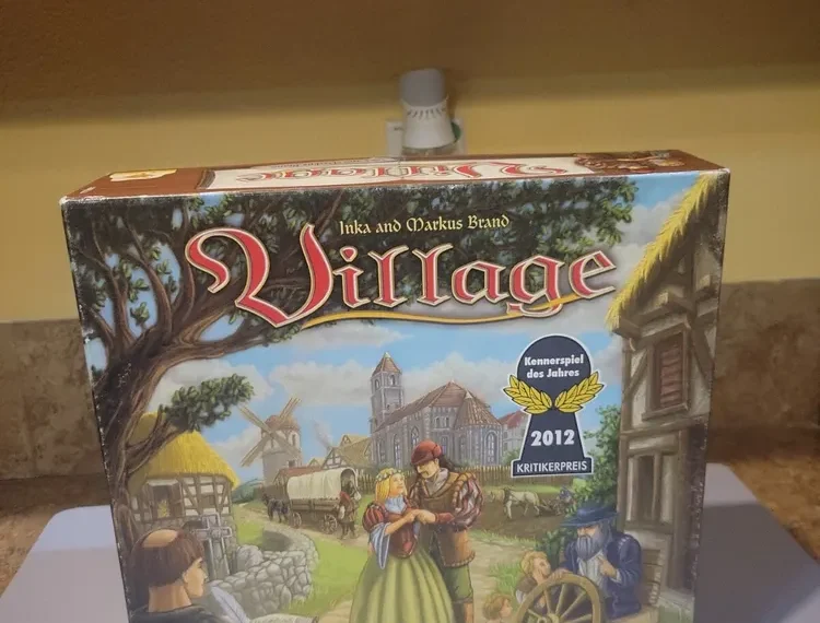 A board game box titled "Village" by Inka and Markus Brand, displayed on a shelf. The box features vivid artwork of medieval village life with characters engaged in various activities and indicates a "2012 Kennerspiel des Jahres Kritikerpreis" award.