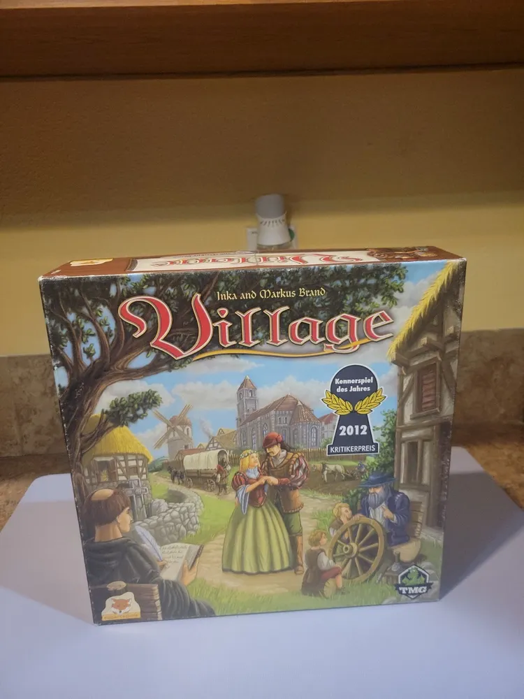 A board game box titled "Village" by Inka and Markus Brand, displayed on a shelf. The box features vivid artwork of medieval village life with characters engaged in various activities and indicates a "2012 Kennerspiel des Jahres Kritikerpreis" award.