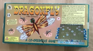 Board game named "Dragonfly" laid on a wooden surface, featuring colorful graphics with a large dragonfly illustration and information about the game's cooperative play nature, suitable for ages 5 to 9, and made from recycled materials.