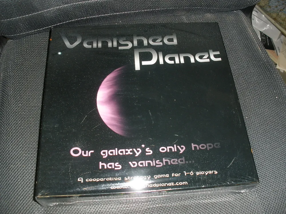 A photo of the boxed board game "Vanished Planet" placed on a dark fabric surface. The box features an image of a planet with the text "Our galaxy's only hope has vanished. A cooperative strategy game for 1-6 players. www.vanishedplanet.com".