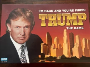 Board game box cover with the title "TRUMP THE GAME" featuring a portrait of a man on the left side and a city skyline on the right, along with the tagline "I'M BACK AND YOU'RE FIRED!" above the title. The Parker Brothers logo and the text "AGE ADULT" are located in the lower left corner.