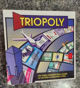 Board game "TRIOPOLY," featuring colorful graphics depicting city landmarks, transportation, and property spaces, with text at the bottom reading "THE THREE-DIMENSIONAL GAME OF BUYING & BUILDING CITIES."