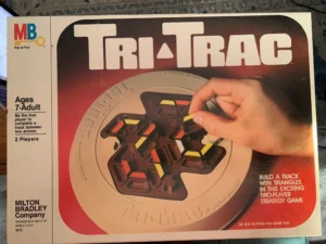 Box cover for the "Tri-Trac" board game by Milton Bradley, showing a hand playing with triangular pieces on a track, with text indicating it is for ages 7 to adult and is a two-player strategy game.