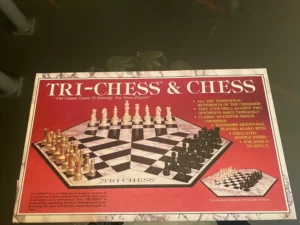 Box cover of TRI-CHESS & CHESS board game featuring a three-player chess set with gold, black, and ivory pieces on a hexagonal board, with text highlighting the game's features such as traditional movements, Staunton design chessmen, and suitability for ages 8 to adult.