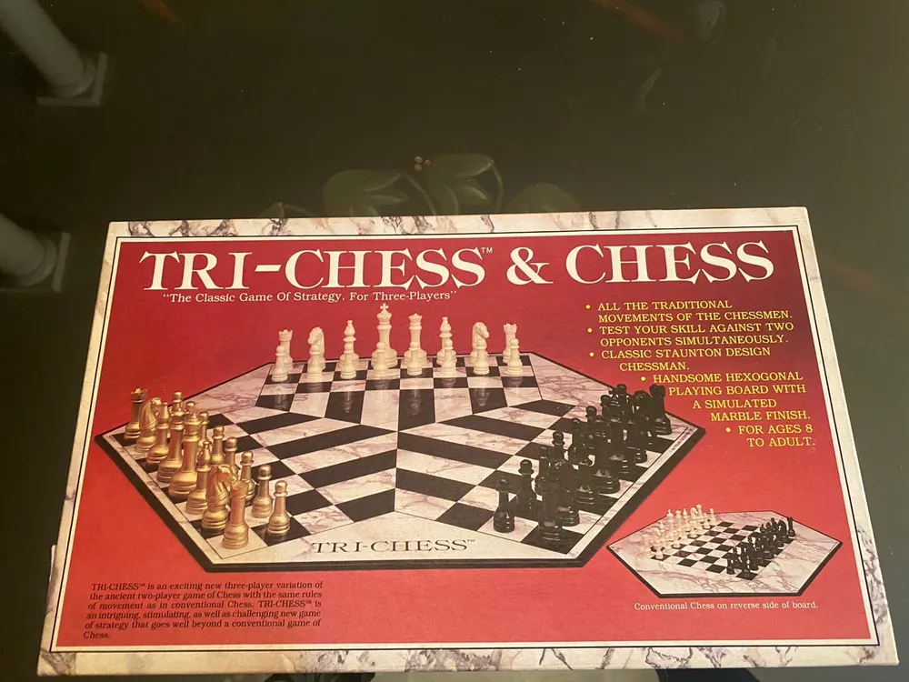 Box cover of TRI-CHESS & CHESS board game featuring a three-player chess set with gold, black, and ivory pieces on a hexagonal board, with text highlighting the game's features such as traditional movements, Staunton design chessmen, and suitability for ages 8 to adult.