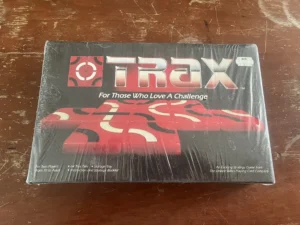 A shrink-wrapped board game named "TRAX" with the tagline "For Those Who Love A Challenge" on a wooden surface. The cover shows a red and black abstract design, and the box has text stating it is for two players ages 10 to adult, includes 64 Trax tiles, a storage tray, and an instruction and strategy booklet. It's noted as an exciting strategy game by The United States Playing Card Company. There is a price tag on the upper right corner with the price "$19.99".