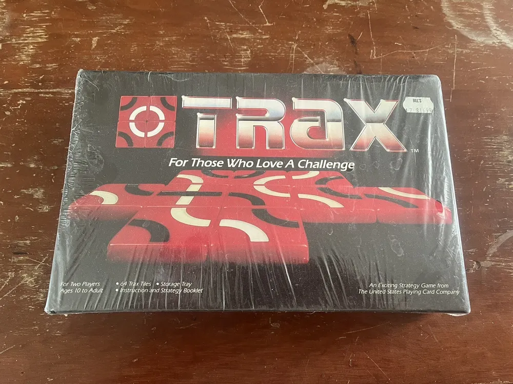 A shrink-wrapped board game named "TRAX" with the tagline "For Those Who Love A Challenge" on a wooden surface. The cover shows a red and black abstract design, and the box has text stating it is for two players ages 10 to adult, includes 64 Trax tiles, a storage tray, and an instruction and strategy booklet. It's noted as an exciting strategy game by The United States Playing Card Company. There is a price tag on the upper right corner with the price "$19.99".