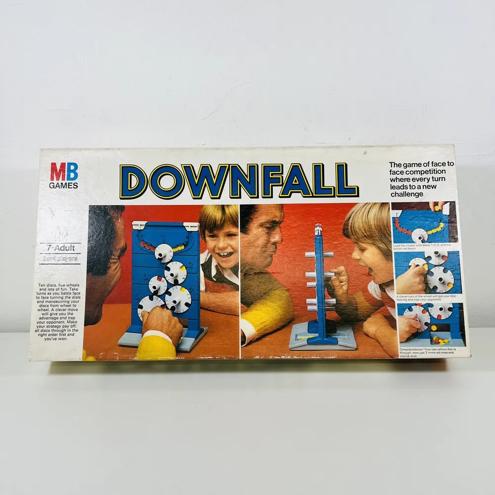 A worn board game box labeled "DOWNFALL" by MB Games, showing images of smiling children playing the game, with close-ups of hands turning knobs on the game mechanism. The text on the box mentions it's for ages 7 to Adult and describes the game as a face to face competition where every turn leads to a new challenge.