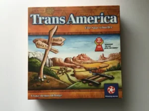 A "TransAmerica" board game box lying on a white surface, featuring a Western American landscape with train tracks and a signpost with city names like "Phoenix", "Santa Fe", "Boston", and "Sacramento". The game is by Franz-Benno Delonge, published by Winning Moves, and has a Spiel des Jahres nomination badge for 2002.