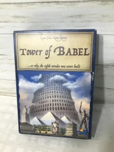 Board game titled "Tower of Babel" by Reiner Knizia displayed on a wooden background. The cover features an illustration of the Tower of Babel among various other wonders of the world, like the Pyramids of Giza and the Colossus of Rhodes.