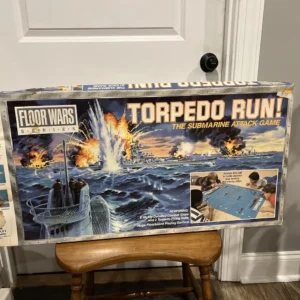 Board game "Torpedo Run! The Submarine Attack Game" from the Floor Wars Series leaning against a wall above a wooden chair. The box art depicts a dramatic naval battle scene with explosions and a submarine in the foreground, along with images of children playing the game. Features are listed including 8 highly detailed combat ships and a huge floorboard playing surface.