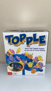 A Topple board game box standing upright on a white surface with the game's name prominent at the top, animated characters balancing on the sides, and an inset photo of three children playing the game, recommended for ages 6 and up.