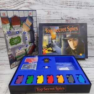 Board game "Top Secret Spies" displayed with its box, components, and game board featuring different color spy pieces and cards on a wooden background.