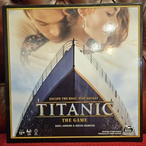 A board game box cover featuring the bow of the Titanic with the title "TITANIC THE GAME" in large golden letters, an image of an embracing couple in the background, and the tagline "Escape the boat, stay afloat". The age recommendation is 12+, and logos for Spin Master Games and creators Daryl Andrews and Adrian Adamescu are also visible, along with a note stating "Knowledge of English Required."