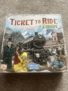 Alt text: The image shows a board game titled "Ticket to Ride Europe" by Alan R. Moon, with illustrations of characters from different social backgrounds and a black steam locomotive set against a mountainous backdrop. The game is produced by Days of Wonder.