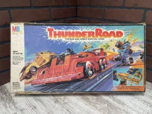 A vintage "Thunder Road" board game box featuring dynamic artwork of vehicles racing and colliding on a road, with a small image showing children playing the game, placed on a brick background.