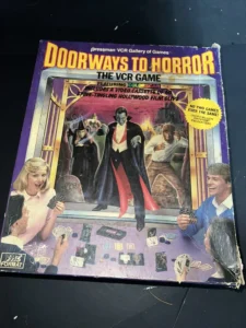 Alt text: A worn-out board game box cover for "Doorways to Horror, The VCR Game," featuring illustrations of a vampire-like character in the center, two players engaged in the game, various game cards spread on the table, and a VHS tape image with the text "Full VHS Format." The box indicates that the game includes a video cassette with 60 minutes of Hollywood film clips, and that no two games are ever the same.