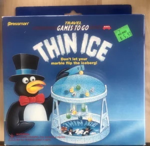 A board game box labeled "TRAVEL GAMES TO GO" with the title "THIN ICE" from Pressman. An illustration of a penguin in a top hat and bow tie is beside a dome-shaped game with marbles on top and the warning "Don't let your marble flip the iceberg!" A Walmart price tag shows $5.97, and a warning label indicates a choking hazard for children under 3 years.