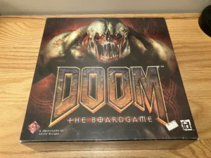 A "DOOM" board game box featuring a prominent graphic of a monstrous creature on a wooden table. The box has the text "THE BOARDGAME" below the title and credits "A BOARDGAME BY KEVIN WILSON" with logos of Fantasy Flight Games and id Software.
