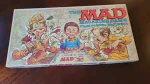 A board game box titled "The MAD Magazine Game from Parker Brothers" with cartoon illustrations of zany characters and currency bills in the background.