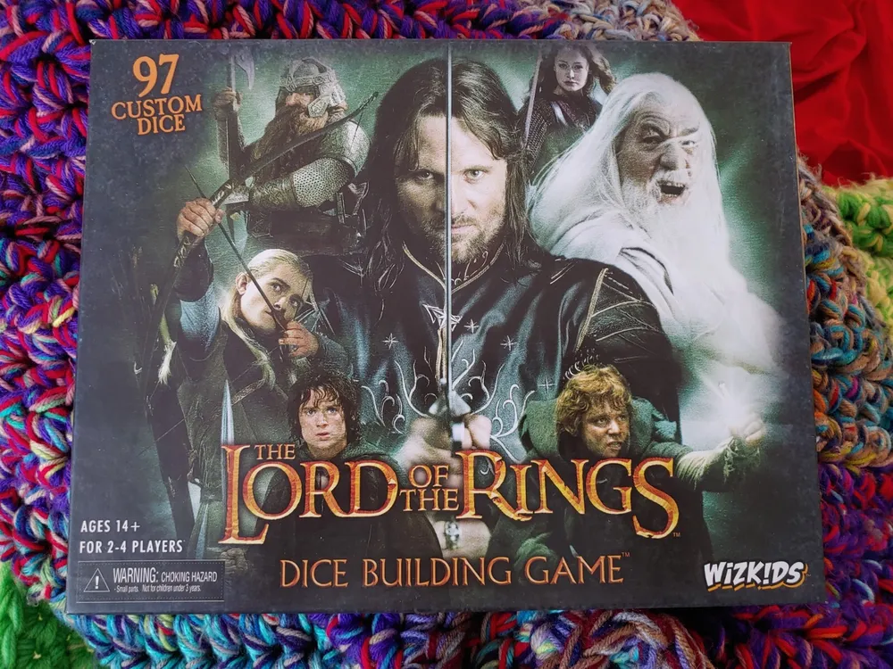 Box cover for "The Lord of the Rings Dice Building Game" by WizKids, showing images of characters from the franchise with "97 Custom Dice" highlighted at the top and an age and player count recommendation at the bottom left corner, all resting on a colorful knit textile.