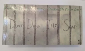 A metal sign with the phrase "Don't Drop the Soap" written across in stylized letters resembling prison bars, with additional text along the top edge reading "Micro to the Macro Effect, Directly Through the Front Door!" and a logo in the bottom right corner.