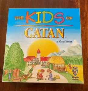 Box cover of "The Kids of Catan" board game showing cartoon-style artwork featuring children engaging in various activities in a colorful medieval village setting, with trees, a rising (or setting) sun in the background, and the game's title and author "Klaus Teuber" at the top. The Mayfair Games logo is visible at the bottom right corner.
