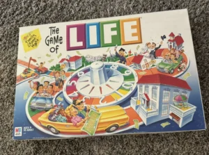 A board game box of "The Game of LIFE" depicting cartoon illustrations of families in cars, houses, and a large spinning wheel, indicating different life stages. The box indicates the game is for ages 9 to adult.