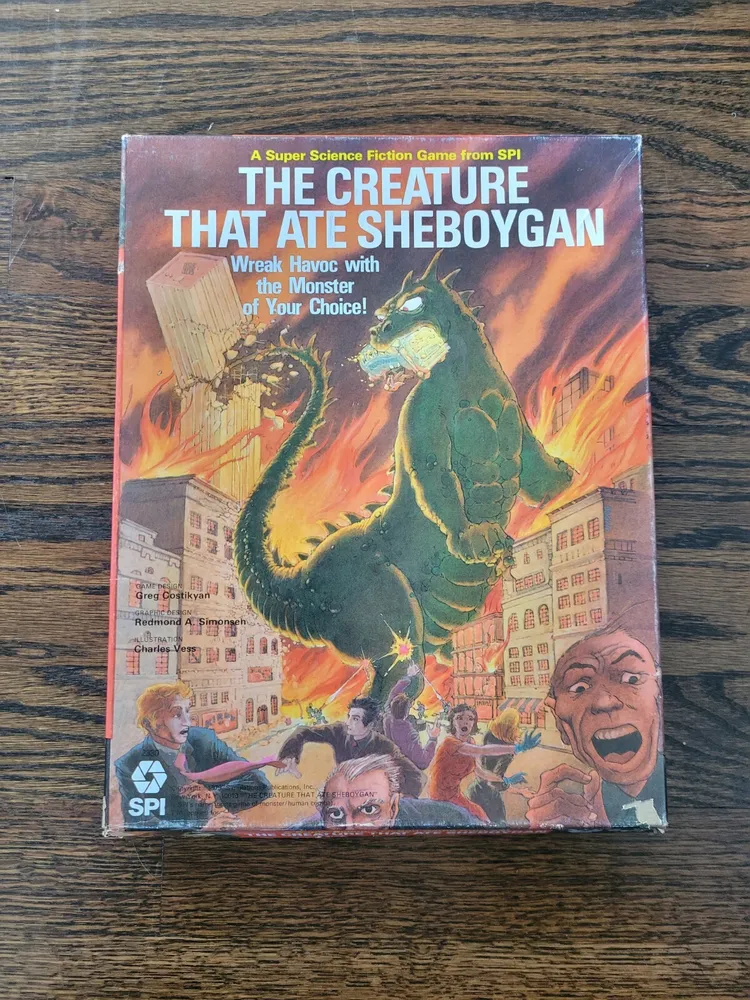Board game cover depicting a giant green monster rampaging through a city with frightened people in the foreground, titled "The Creature That Ate Sheboygan," placed on a wooden surface.