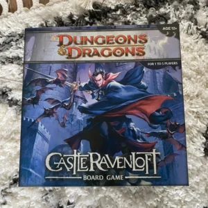 A "Dungeons & Dragons Castle Ravenloft" board game box lying on a textured white carpet. The cover features an illustration of a vampire and flying bats with gothic lettering. The game is labeled for ages 12+ and for 1 to 5 players.