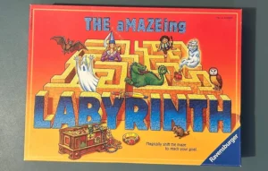Board game "The aMAZEing Labyrinth" cover with colorful illustrations of fantasy characters and a maze, along with the text "Magically shift the maze to reach your goal" and the Ravensburger logo.