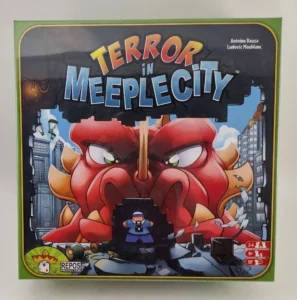 Board game box cover for "Terror in Meeple City" showing a stylized, angry red monster destroying a cityscape, with game details and logos around the edges.