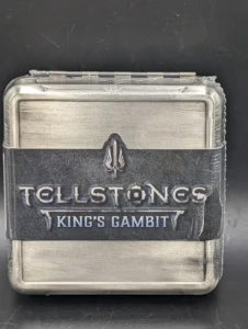 A metal tin with the embossed text "TELLSTONES KING'S GAMBIT" secured with a black rubber band, against a black background.