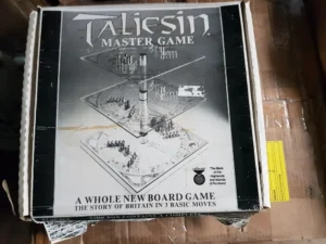 Alt text: A worn-out box of the "Taliesin Master Game" on a wooden surface with the tagline "A WHOLE NEW BOARD GAME - THE STORY OF BRITAIN IN 3 BASIC MOVES," showing an illustration of the game board and pieces with text indicating it is "craftmade" and "The Mark of the Highlands of Scotland." The box is opened and slightly damaged, with clear packaging tape on the corner and a yellow sticker on the side.
