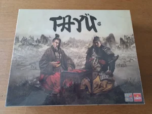 Box cover for a board game featuring two historical Asian figures playing a strategy game, with a detailed ancient battlefield in the background, the game's title in large Asian characters at the top, and logos indicating the number of players, age suitability, and the manufacturer Goliath at the bottom right corner.