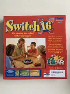 A board game box titled "Switch 16" by TOMY for ages 7 and up, featuring an image of two adults and two children engaging happily in gameplay around a table. The box contains a description of the game, which is a dice-rolling, card-swapping game, along with advertisement for other TOMY games like "Rummikub" at the bottom. The box also displays the CE mark and a barcode.
