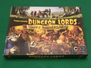 The image shows the board game "Dungeon Lords: Happy Anniversary" by Vlaada Chvátil. The game box features colorful fantasy artwork with various creatures and characters celebrating, with a prominent figure of a smiling creature in the foreground. The logo of Czech Games Edition is visible in the bottom right corner. The box is resting on a green surface.