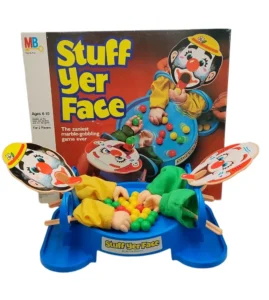 A vintage "Stuff Yer Face" board game by Milton Bradley displayed in front of its box, featuring two clown face paddles, a blue circular base with hands holding multicolored marbles, and green clown pants serving as marble storage.