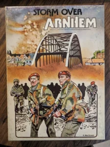 Alt text: A worn board game cover for "Storm Over Arnhem" depicting soldiers in combat gear with a prominent bridge in the background, suggestive of a wartime theme.