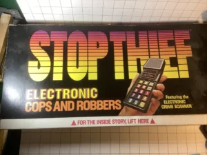 A vintage board game box with the title "STOP THIEF" in bold gradient letters, subtitled "ELECTRONIC COPS AND ROBBERS" with an image of a hand holding a device labeled as "ELECTRONIC CRIME SCANNER" and text at the bottom that says "Featuring the ELECTRONIC CRIME SCANNER" and "FOR THE INSIDE STORY, LIFT HERE" with an arrow pointing downwards.