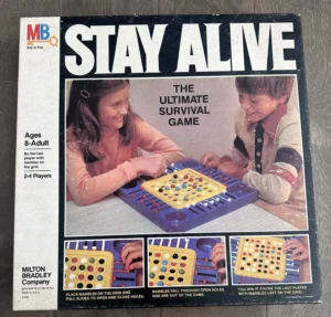 Box cover of the "Stay Alive" board game by Milton Bradley showing two children playing the game with the tagline "THE ULTIMATE SURVIVAL GAME" and instructions for ages 8-Adult, 2-4 players, including gameplay images on the lower side.
